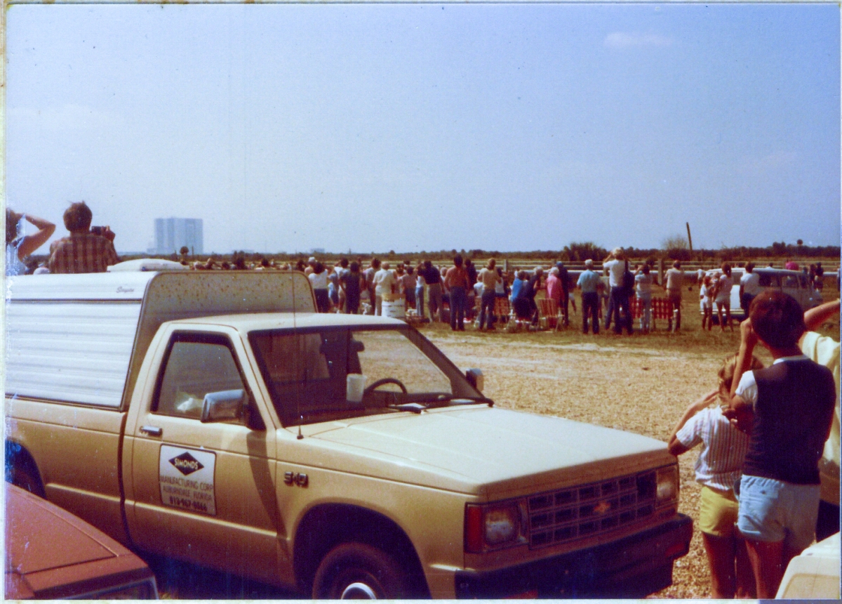 Employees and their families watch as the Space Shuttle, which is sensibly invisible in this image, pre-flares mere seconds prior to touchdown on runway 33 at the Kennedy Space Center, in Florida. In the distance, the Vertical Assembly Building, where Apollo Program rockets and then the Space Shuttles, were assembled before being rolled out to their launch pads. This view is taken from just south of the main gate at Pad 39-B, looking southwest, right next to the crawlerway, a very small part of which can be seen just beyond the hood of the small pickup truck in the foreground.